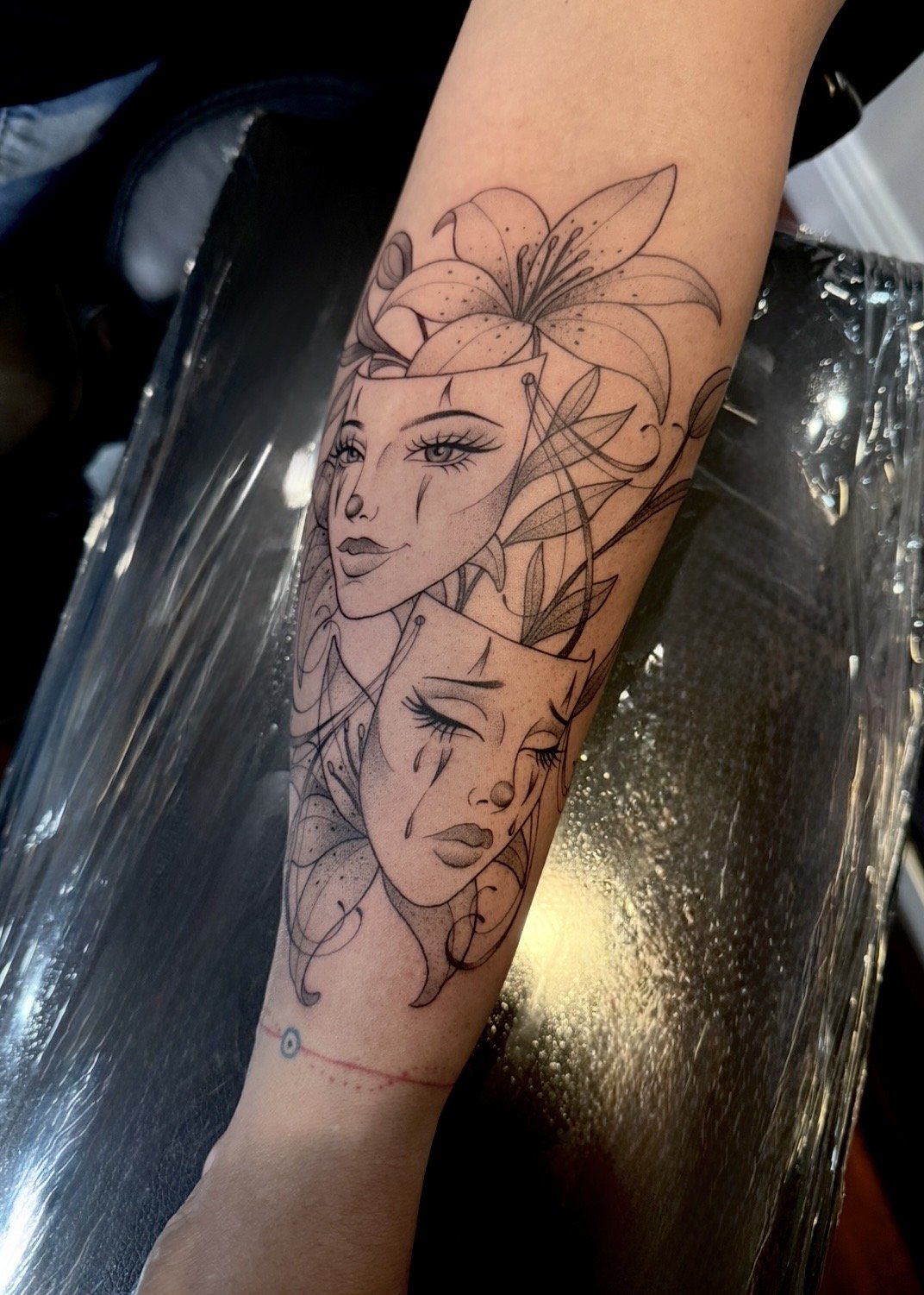 Fine line comedy and tragedy masks with lilies forearm tattoo by Evelyn Garcia